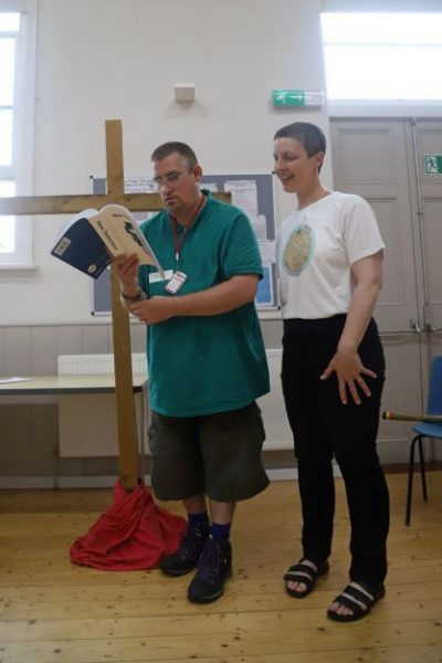 A man stands reading aloud from the accessible Bible, in front of a person-sized wooden cross,. A woman, wearing a t-shirt showing she's a helper at the group, stands next to him