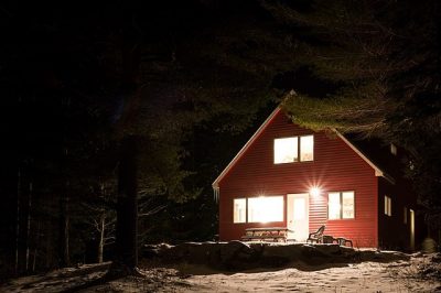 A cosy-looking home with lights on, surrounded by a snowy field and hedge