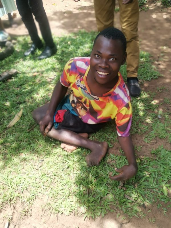 Ugandan man with mis-shaped legs tucked under him, sitting on the ground, smiling.