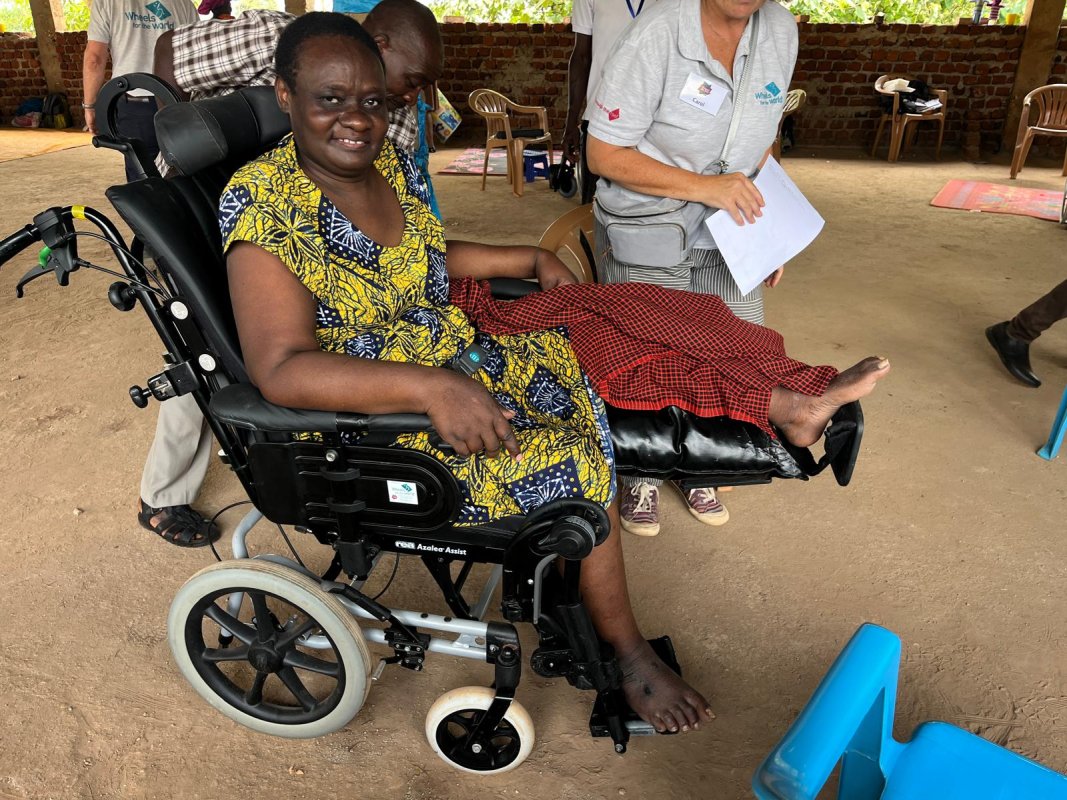 A disabled Ugandan woman in a yellow dress, sat in a wheelchair, with her leg supported out in front of her. 