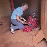 An elderly Ugandan lady, smiling, sat on the floor in a mud hut with a female UK volunteer.