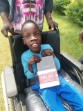 A Ugandan boy, smiling, sat in a wheelchair, holding a Bible