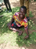 Ugandan man with mis-shaped legs tucked under him, sitting on the ground, smiling.