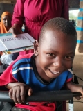 A young Ugandan boy, smiling,  holding a walking frame.