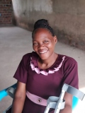 A young Ugandan lady in a purple dress, smiling, sat in a chair, holding crutches.