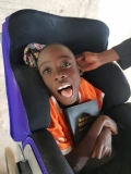 A Ugandan boy with mis-shaped limbs, smiling, sat in a wheelchair, holding a Bible
