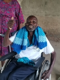 A Ugandan man with a beard, sat in a wheelchair, doing a 'thumbs up' sign.