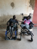 Two disabled Ugandan people, a man and a woman, sat in wheelchairs in a concrete building.