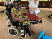 A disabled Ugandan woman in a yellow dress, sat in a wheelchair, with her leg supported out in front of her. 