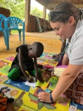 A young Ugandan boy playing on a coloured mat, next to a female UK volunteer.
