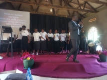 Ugandan church stage with a choir wearing black and white. and a man in a suit with a microphone.