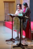 Blind Indian lady in a sari, reading from a braille Bible (from John 9), stood at the front of a hall, with another Indian lady holding a microphone for her.