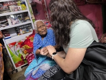 A grey-haired Indian lady sat facing a young female UK volunteer with long brown wavy hair. They are holding hands.