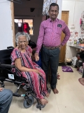 A smiling older Indian woman in a wheelchair, wearing a pink, purple and white striped dress, with an Indian man in a pink striped shirt stood beside her.