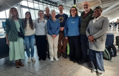 Group photo of 10 people at  a UK airport, smiling, with suitcases. 