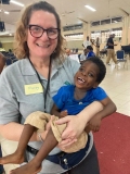 A smiling woman wearing glasses, carrying a small Ghanain boy in a blue T shirt.
