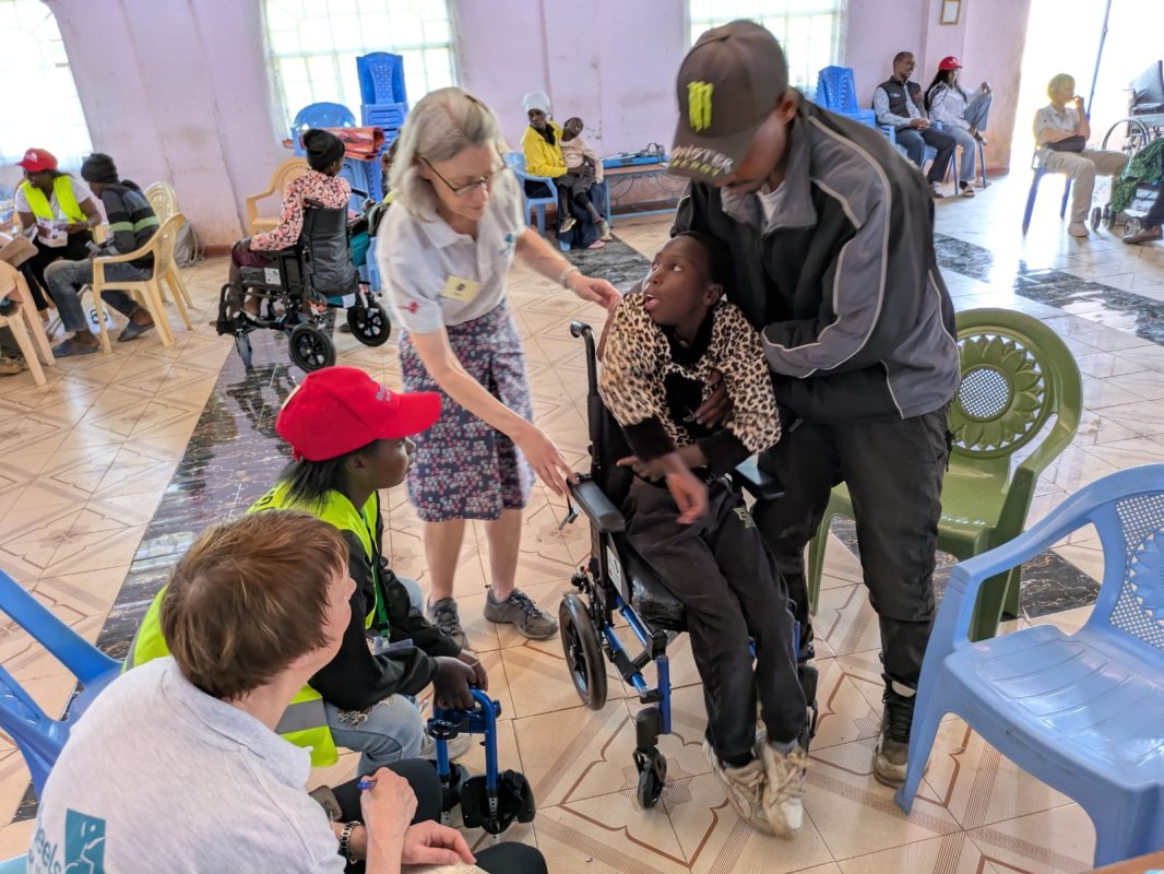 A Kenyan father lifts his disabled daughter, who is wearing a leopard-print-top, into a wheelchair, as 3 volunteers watch or help