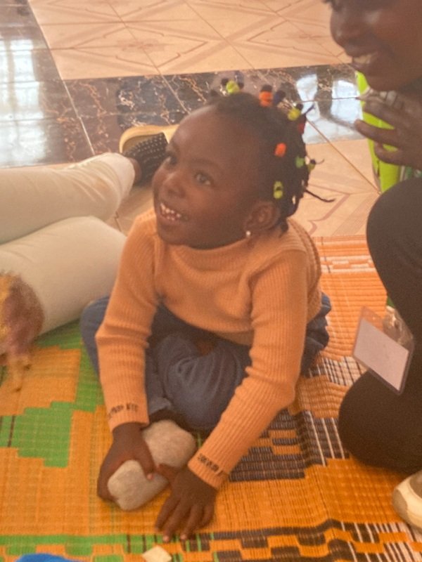 Josephine B, a 5 year old Kenya girl, smiling, sat on floor with local volunteers