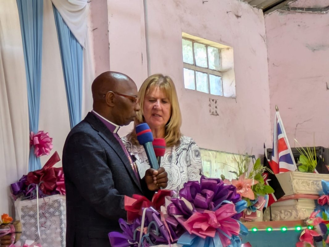 Kathy, a fair haired female UK volunteer, and Pastor Davis, a Kenyan minister wearing glasses: speaking at the front of a Church
