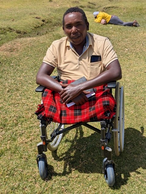 A Kenyan man, sat in a wheelchair outdoors, with a tartan blanket over his knees; you can see that he has no legs below the knee: he is a double amputee.