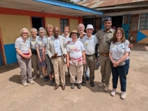 A group of ten UK volunteers stood outside in a courtyard in Kenya, with the local pastor (Davis) and a local military leader. in uniform