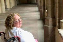 Sarah sits in her wheelchair in a corridor in an abbey or church. She's looking back over her shoulder at the camera.