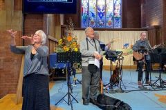 Worship time at our 25th anniversary event. Mike, our chairman, stands singing, with his assistance dog lying at him feet. To the side, a BSL interpreter is signing words to the song, and the band, led by Graham Kendrick, is playing a praise song.