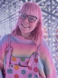 Triona Brading - a smiling young woman with glasses and shoulder-length pink hair, wearing a pink dungarees with spots on,.