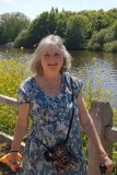 Karen is standing in front of a fenced-off lake, wearing a blue top and using a walking stick