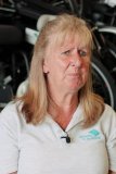 Kathy has blonde hair and is wearing a wheels for the world t-shirt. She's sitting in front of some stacked refurbished wheelchairs. 