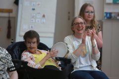 A group of people smiling and laughing - one lady in a wheelchair is playing a tambourine, one lady is clapping, and a young girl  is standing next to them, watching. Behind them is a white board with a visual timetable showing symbols of clock times and activities.