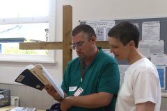 Close-up photo of a man and a woman standing in a church hall, next to a large wooden cross. The man is reading the  Bible and the woman is encouraging and supporting him with the reading.