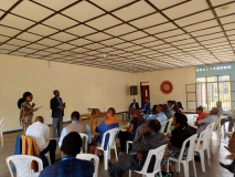 A large group of East African people sat in rows of plastic chairs, listening to two speakers - a man and a woman.