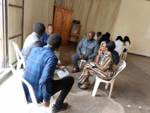 Two groups of about six East African people in each, sat in circles on plastic chairs in a hall.