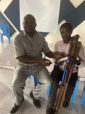 A Rwandan man sat on a chair, presenting an audio Bible to another seated Rwandan  man holding crutches.