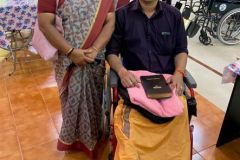 An Indian couple -- a man is sat in his new wheelchair, with a Bible on his lap, and a woman is standing next to him