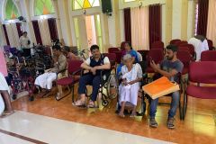 A group of local people wait for adjustments to their wheelchairs. They're chatting, and relaxing by some chairs in a large hall space.